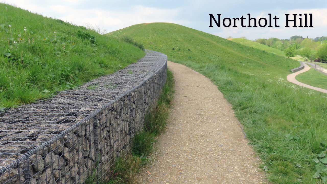Northolt Hill walking path in Northala Fields, West London. Wide view of a gravel footpath and stone gabion wall leading up the grassy mound at Northolt Hill, Northala Fields, London, UK, on a cloudy day.