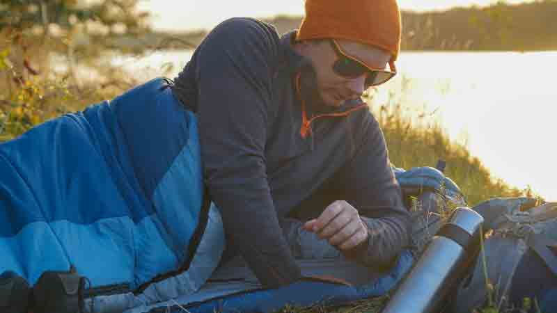 Camper relaxing in a sleeping bag by the water in early morning light Person using a sleeping bag by a lakeside at sunrise during a camping trip