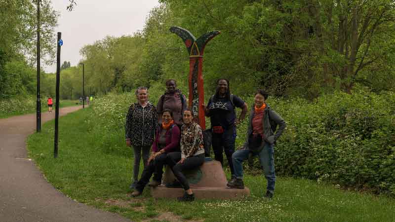 Spartan hikers from Hiking in London Club on Waterlink Way trail, South London Group of women hikers from Hiking in London Club posing next to a red and green trail sign on Waterlink Way, surrounded by greenery and trees.