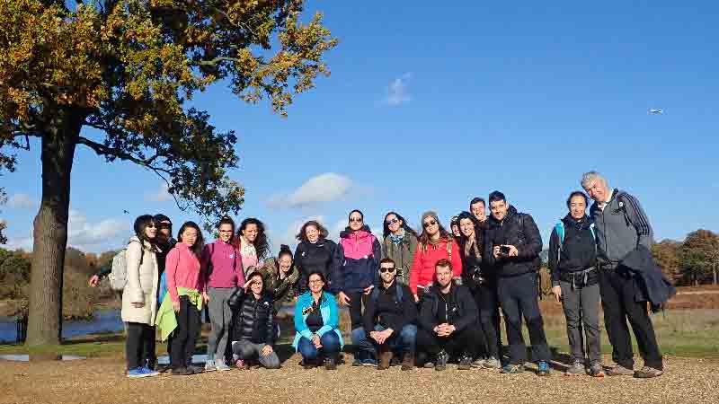 Group of hikers posing by a lake under an oak tree at Wimbledon Park, blue sky and plane in the distance.