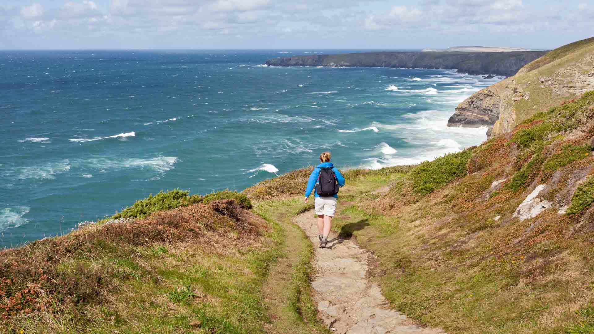 Hiker walking along coastal path near cliffs and ocean in Cornwall, UK.