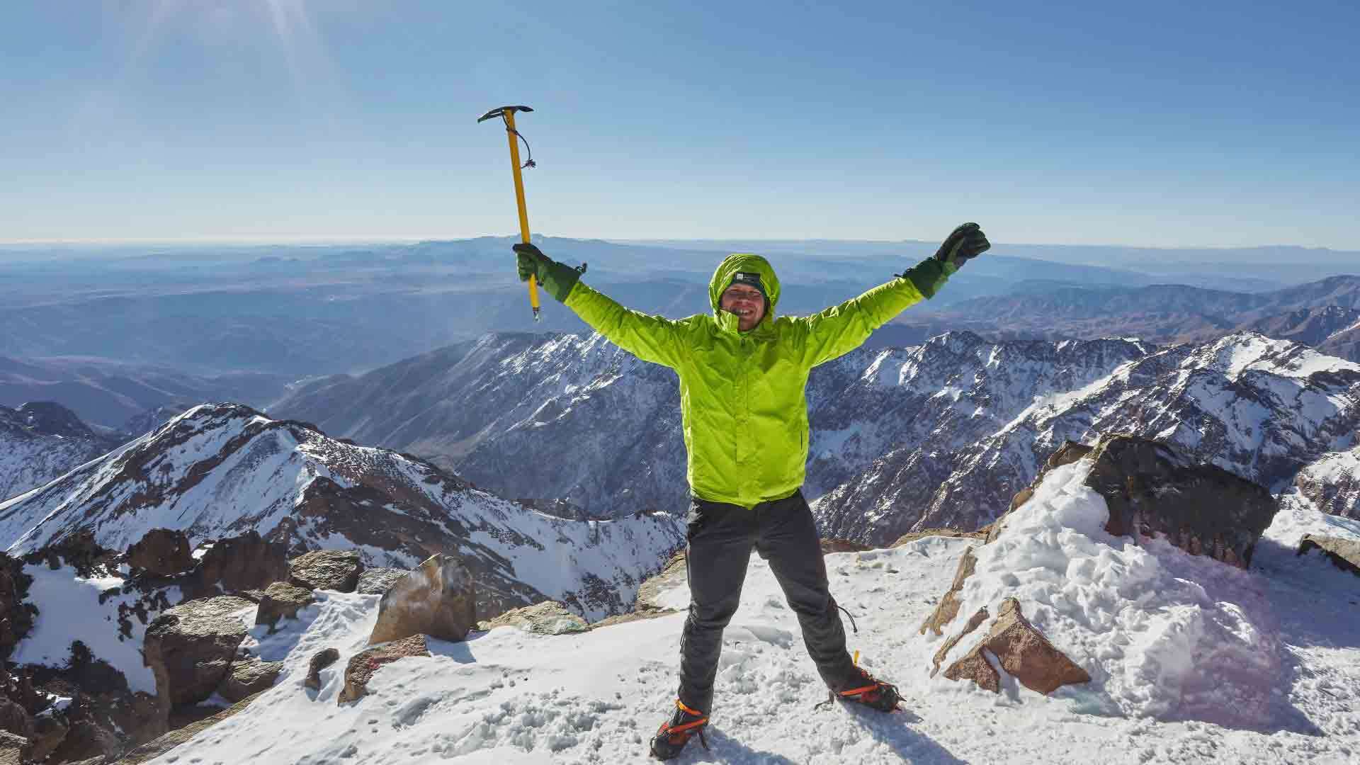 Winter ascent of Mount Toubkal – Climber at the summit with ice axe Climber on snowy summit of Mount Toubkal in winter, wearing bright green jacket and holding an ice axe