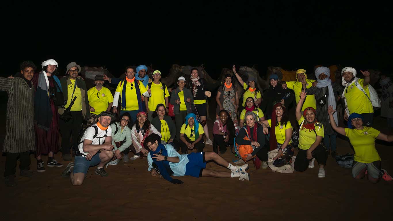 Tour group in the Sahara Desert at night – “Morocco 6-Day Desert: Marrakesh & Beyond” adventure. Large group of travelers in bright clothing kneeling and standing beside camels at night on the Sahara Desert during a six-day Morocco tour from Marrakesh.
