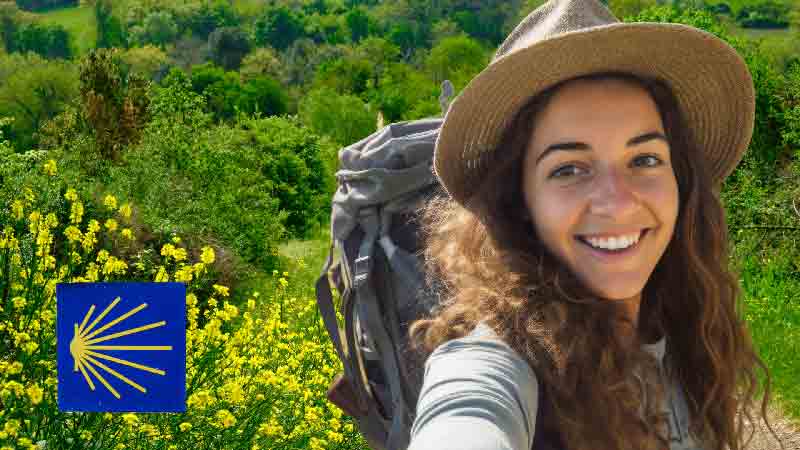 Pilgrim enjoying the Camino Inglés near Sigüeiro, Galicia Smiling female pilgrim on the English Way in Galicia, surrounded by green fields and yellow wildflowers, with a Camino de Santiago shell sign visible.