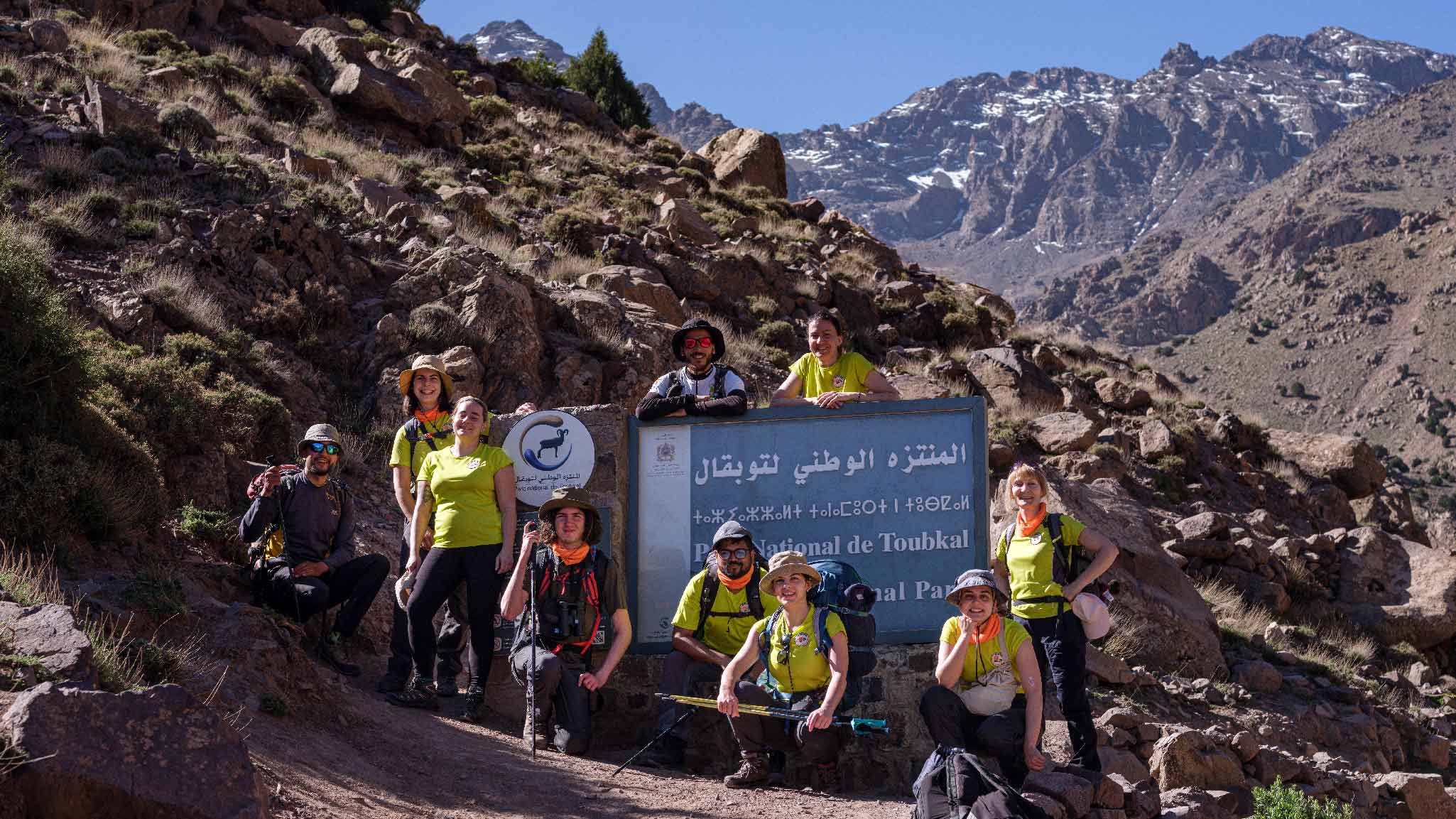 Hikers at Toubkal National Park entrance sign A group of eight hikers in bright shirts and backpacks standing and kneeling around a blue “Parc National de Toubkal” sign, with rocky slopes and snow-dusted peaks behind them