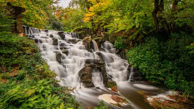Cascading Waterfall at Virginia Water – A Scenic Highlight on the Circular Trail. Cascading waterfall surrounded by lush greenery at Virginia Water Circular trail.